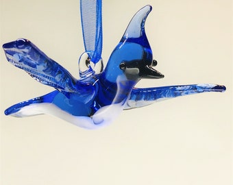 Op de afbeelding: Een blauwe glazen ornament in de vorm van een blauwe gaaien met een witte buik. De ornament heeft een zilveren lus om op te hangen.