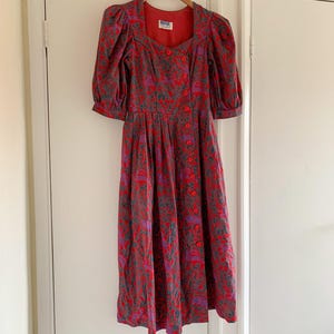 Könnte beinhalten: Ein rotes und violettes Blumenkleid mit quadratischem Ausschnitt und Knopfleiste. Das Kleid hat Puffärmel und eine Midi-Länge.