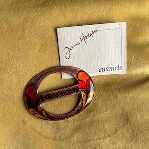 Op de afbeelding: Ovale, bruine emaille gesp met een bloemmotief in rood, oranje, geel en zwart. De gesp ligt naast een kaart met de naam "Janna Hodgson" en het woord "email".