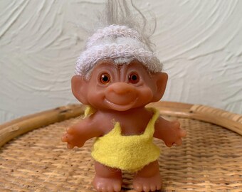 May include: A small, brown troll doll wearing a white knitted hat and a yellow swimsuit. The doll has brown hair and orange eyes.