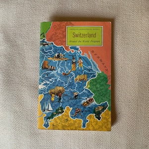 以下が含まれることがあります： 赤、白、青のカラーリングが施された、スイスのヴィンテージイラストマップ。地図には、国の主要都市、湖、山々が示されています。表紙には、「アメリカ地理学会」と「世界一周プログラム」の文字が印刷されています。