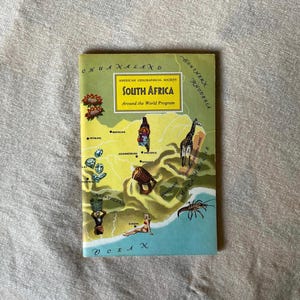 Op de afbeelding: Een vintage reisgids getiteld "Zuid-Afrika" van de American Geographical Society's "Around the World Program". De omslag toont een kaart van Zuid-Afrika met illustraties van een giraffe, een vrouw en andere culturele elementen. Het kleurenpalet omvat geel, groen en blauw.