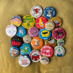 May include: A selection of vintage pin badges with diverse designs and text. The badges showcase illustrations of animals, book-related themes, and phrases such as "Haunt Your Library" and "Libraries are Magic". The badges are round and colourful.