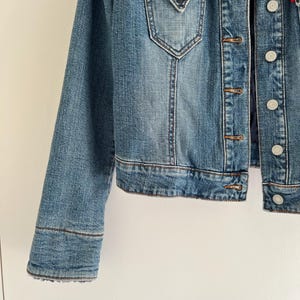May include: A medium-wash denim jacket with a button-up front, two chest pockets, and a dark blue faux fur collar. The jacket features copper-colored buttons and a red logo tag.