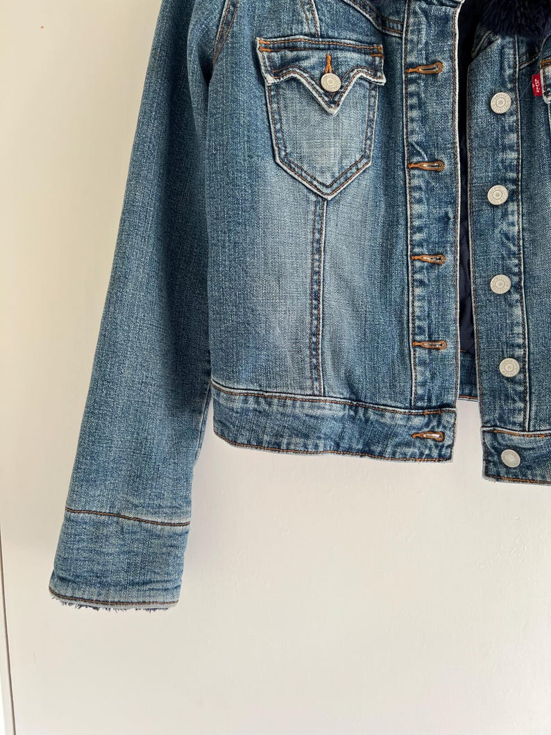 May include: A medium-wash denim jacket with a button-up front, two chest pockets, and a dark blue faux fur collar. The jacket features copper-colored buttons and a red logo tag.