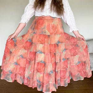 May include: A long, tiered skirt featuring a floral print in peach, coral, and blue hues. The skirt is made of a light fabric, with the floral design repeated across the layers. It is worn with a white blouse, creating a bohemian aesthetic.