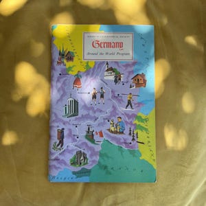 Op de afbeelding: Een vintage boekomslag getiteld "Germany Around the World Program" van de American Geographical Society. De omslag toont een kaart van Duitsland met illustraties van mensen en bezienswaardigheden. Het kleurenpalet omvat blauw, groen, geel en paars.