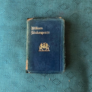 以下が含まれることがあります： 表紙に金色の文字でWilliam Shakespeareと刻印された、小さな濃紺のハードカバー本。文字の下には、中央の盾を挟むように2頭の紋章獣が描かれた金色のエンブレムがある。本の縁には摩耗が見られる。