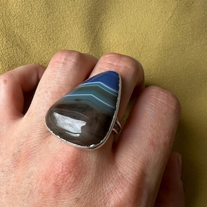 May include: A large statement ring with a polished, triangular agate stone. The stone has bands of blue, teal, and brown, set in a silver-coloured metal band. The ring is worn on a finger.