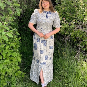 May include: A white maxi dress with a blue floral pattern. The dress has a high neck, puffed sleeves, and a tie at the neck. The skirt is a patchwork of blue and white squares.