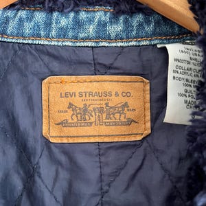 May include: Close-up of the inside of a denim jacket. The jacket features a navy blue quilted lining and a brown leather label with the Levi Strauss & Co. logo. The jacket is trimmed with a dark blue, fuzzy material.