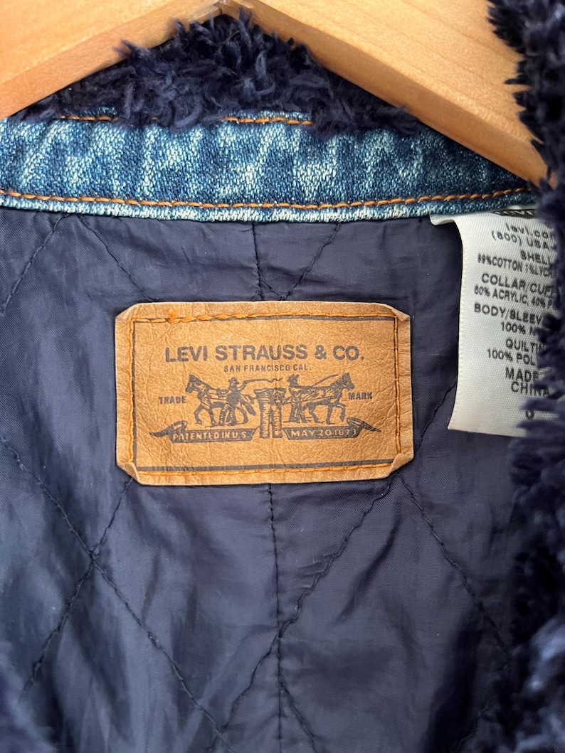 May include: Close-up of the inside of a denim jacket. The jacket features a navy blue quilted lining and a brown leather label with the Levi Strauss & Co. logo. The jacket is trimmed with a dark blue, fuzzy material.