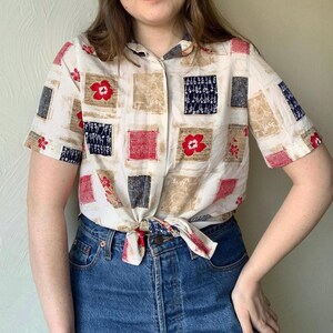 Op de afbeelding: Een wit, kort mouwig overhemd met knopen en een geometrisch patroon in tinten rood, blauw en bruin. Het overhemd is in de taille geknoopt en wordt gedragen met een blauwe jeans.