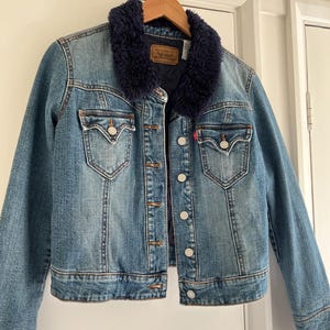 May include: A blue denim jacket with a dark blue faux fur collar. The jacket has two chest pockets, button closures, and a red Levi's tag. It is hanging on a wooden hanger.