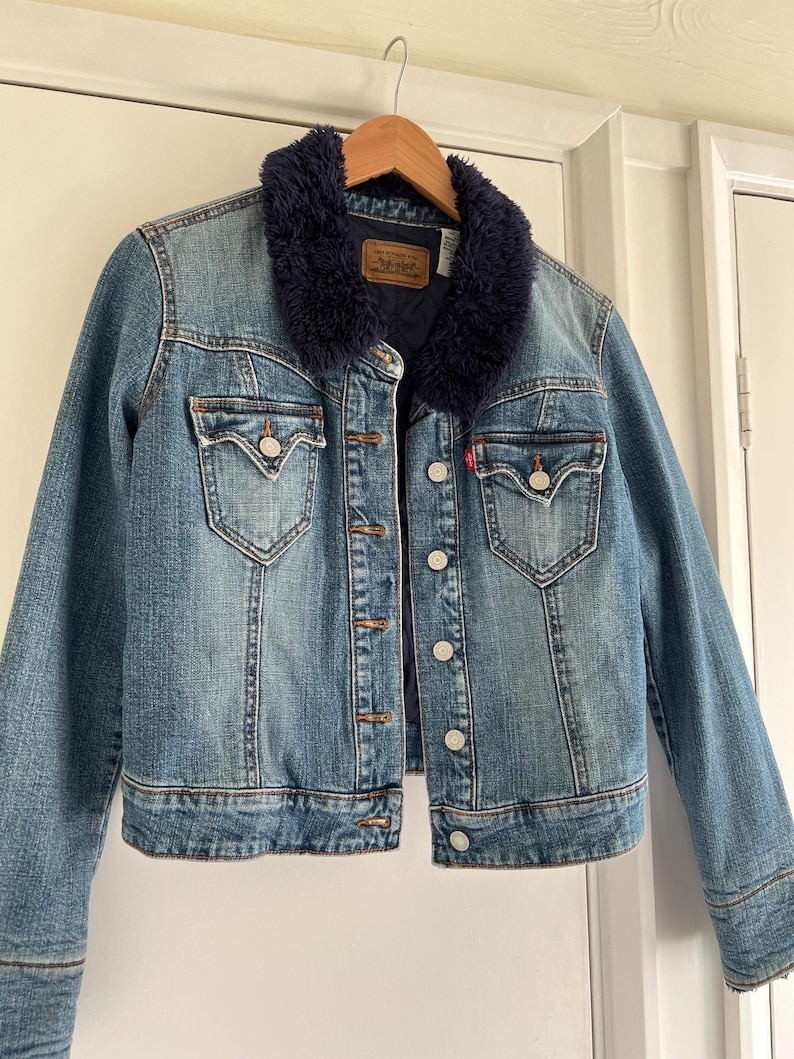 May include: A blue denim jacket with a dark blue faux fur collar. The jacket has two chest pockets, button closures, and a red Levi's tag. It is hanging on a wooden hanger.