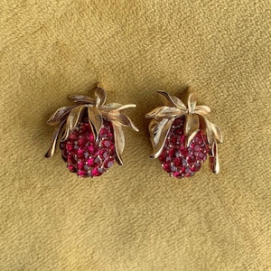 May include: A pair of gold tone earrings shaped like strawberries with red rhinestone accents.