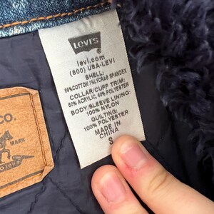 May include: Close-up of a Levi's label on a denim jacket. The label includes the Levi's logo, website, and fabric composition details. The jacket has a blue quilted lining and a small size tag.