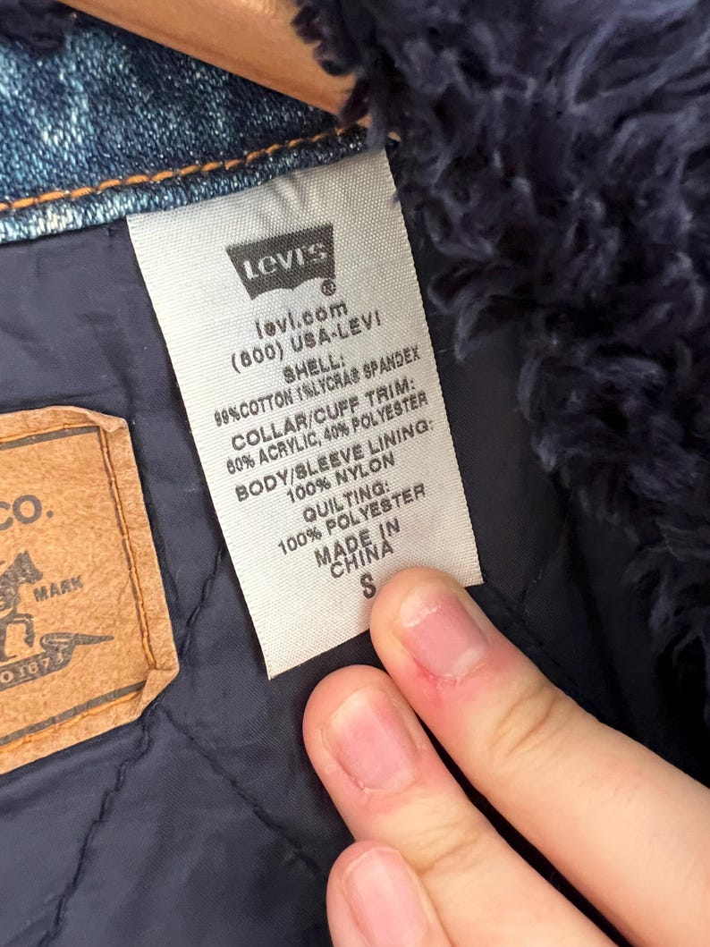 May include: Close-up of a Levi's label on a denim jacket. The label includes the Levi's logo, website, and fabric composition details. The jacket has a blue quilted lining and a small size tag.