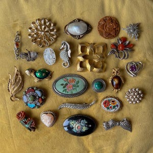 May include: A collection of vintage brooches in various shapes and designs, including floral, geometric, and animal motifs. The brooches feature a range of materials, such as metal, gemstones, and mother-of-pearl, displayed on a yellow background.
