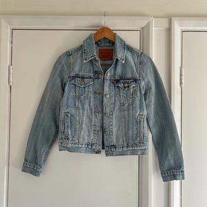 Puede incluir: Chaqueta vaquera azul claro con diseño clásico. Cuello camisero, cierre de botones y dos bolsillos en el pecho. La chaqueta tiene un lavado descolorido y una etiqueta roja en el bolsillo del pecho. Cuelga de una percha de madera.