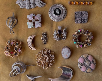 May include: A collection of vintage brooches in various shapes and designs. The brooches include floral, geometric, and figural styles, with materials like metal, rhinestones, and mother-of-pearl. The color palette includes silver, gold, and iridescent hues.