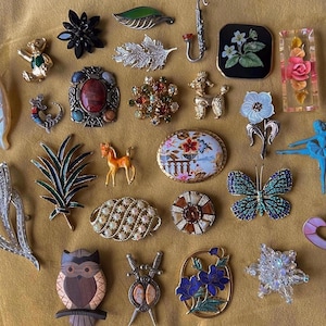 May include: A collection of vintage brooches and pins in various shapes and colors. Includes a butterfly, floral, and animal designs. The brooches are made of metal, enamel, and rhinestones. The collection is displayed on a golden-yellow surface.