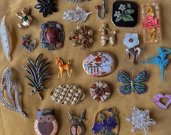 May include: A collection of vintage brooches and pins in various shapes and colors. Includes a butterfly, floral, and animal designs. The brooches are made of metal, enamel, and rhinestones. The collection is displayed on a golden-yellow surface.