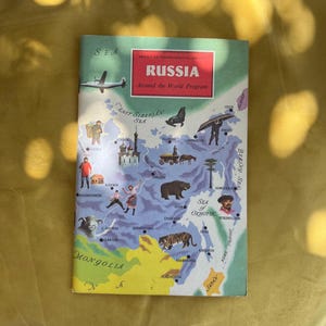 Op de afbeelding: Een vintage boekomslag getiteld "RUSSIA Around the World Program" van de American Geographical Society. De omslag toont een kaart van Rusland met illustraties van dieren, mensen en bezienswaardigheden. Het kleurenpalet omvat tinten blauw, groen, geel en rood.