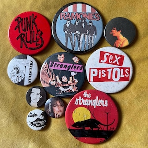 May include: A collection of ten circular pin-back buttons featuring various punk rock band imagery and text. Designs include the Ramones, Sex Pistols, and The Stranglers, with graphics such as a rat, band portraits, and the text Punk Rules.