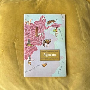 May include: A vintage book cover titled "Afghanistan" from the American Geographical Society's "Around the World Program." The cover features a map of Afghanistan with illustrations of animals, people, and cultural elements in a colourful design.