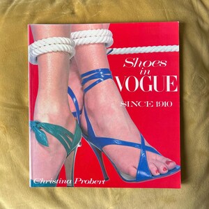 May include: A red book cover with the title "Shoes in VOGUE SINCE 1910" features a photo of feet wearing blue and green high-heeled sandals. The book is by Christina Probert. The feet are adorned with white rope bracelets.