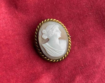 May include: A gold-toned cameo brooch featuring a white cameo carving of a woman's profile. The cameo is set in a rope-like border.