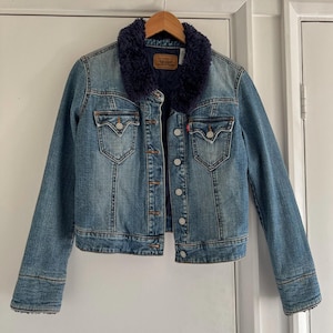 May include: A blue denim jacket with a dark blue faux fur collar. The jacket has a button-up front, two chest pockets, and two side pockets. The Levi's brand label is visible on the collar. The jacket is hanging on a wooden hanger.