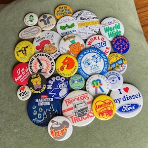 May include: A collection of vintage pinback buttons with various designs and text. The buttons feature logos, illustrations, and phrases, including "I love my diesel" and "Haunted House." The buttons are in various colors and sizes, and are scattered on a green surface.
