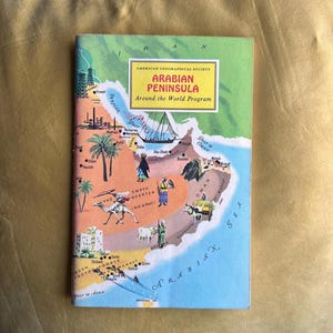 Op de afbeelding: Een vintage boek getiteld "Arabian Peninsula" van de American Geographical Society's "Around the World Program". De omslag toont een kleurrijke kaart met illustraties van kamelen, boten en palmbomen, met tekst in een gele rechthoek.