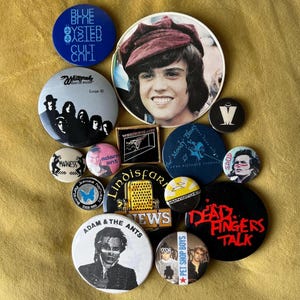 May include: A collection of vintage band and music-themed buttons and pins. The pins feature images of musicians, band logos, and text, including "Adam & The Ants", "Pet Shop Boys", and "Dead Fingers Talk". The buttons are in various shapes and sizes.
