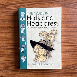 May include: A book titled "The Mode in Hats and Headdress" with illustrations of various hats and headwear. The cover features a woman wearing a large black hat with feathers. The book is on a wooden surface.