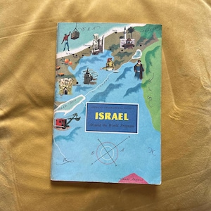 Könnte beinhalten: Ein Vintage-Buch mit dem Titel "ISRAEL Around the World Program" der American Geographical Society. Das Cover zeigt eine Karte von Israel mit Illustrationen von Sehenswürdigkeiten und Figuren. Das Buch hat einen blauen Einband mit einem farbenfrohen Kartendesign.
