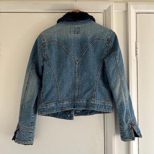 May include: A light blue denim jacket with a dark blue faux fur collar. The jacket has a classic design with button closures on the cuffs and front. The back features a unique yoke detail.