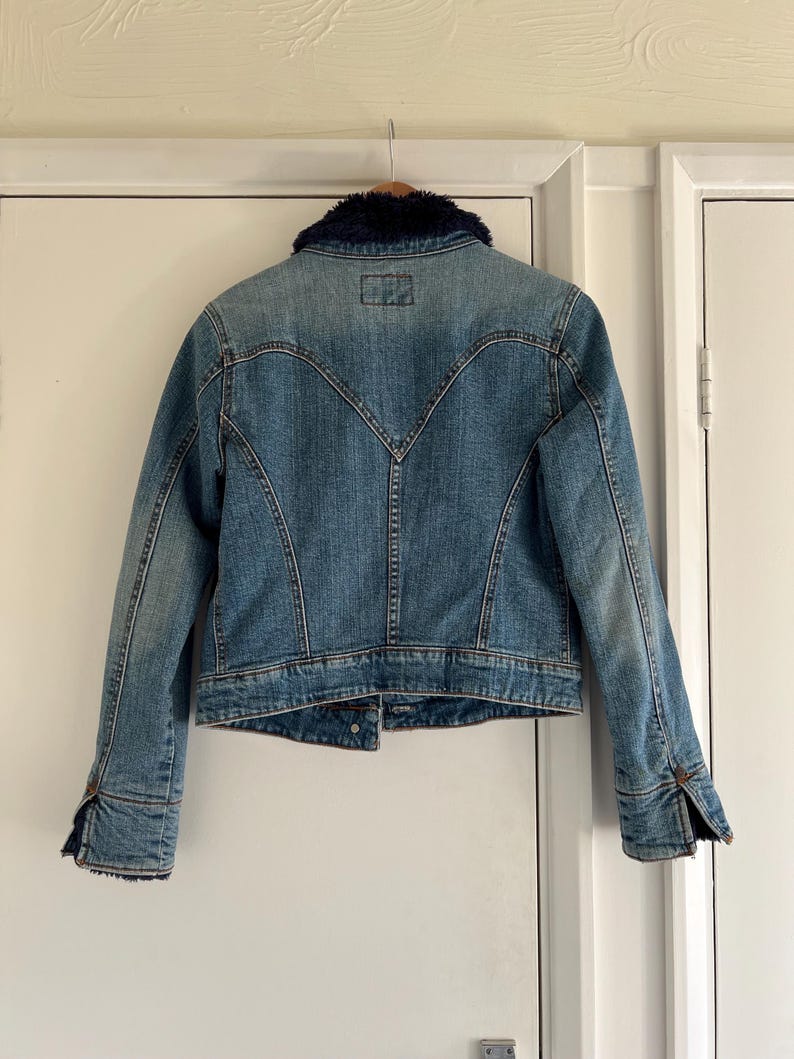 May include: A light blue denim jacket with a dark blue faux fur collar. The jacket has a classic design with button closures on the cuffs and front. The back features a unique yoke detail.