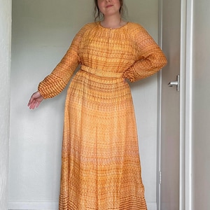 May include: A long orange dress with a textured pattern. The dress features long sleeves, a round neckline, and a fitted waist. The skirt flows to the floor. The fabric appears lightweight. The dress is a warm orange colour with darker orange detailing.