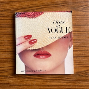 May include: A book cover showing a woman's face with red lipstick and nails, partly hidden by a straw hat. The title "Hats in Vogue Since 1910" is visible, with the author's name, Christina Probert, at the bottom.