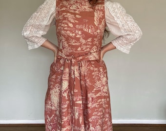 May include: A rust-colored dress with a cream-colored toile pattern featuring a tied waist and a high neckline. The dress has a sleeveless bodice and is paired with white lace sleeves.