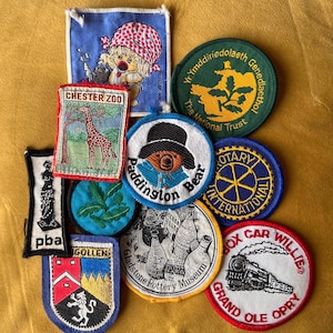 Può includere: Una collezione di toppe ricamate con vari disegni. Include una toppa con una giraffa e il testo "CHESTER ZOO", una toppa di Paddington Bear e una toppa "BOX CAR WILLIE" con un'illustrazione di un treno. Altre toppe presentano testo e loghi.