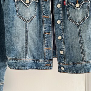 May include: A blue denim jacket with a dark blue faux fur collar. The jacket has two front pockets with button closures, a button-up front, and a small red tag. The jacket is a medium wash denim.