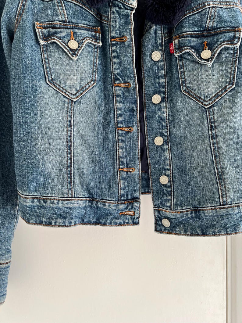 May include: A blue denim jacket with a dark blue faux fur collar. The jacket has two front pockets with button closures, a button-up front, and a small red tag. The jacket is a medium wash denim.