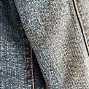 May include: Close-up of a denim jacket sleeve. The blue fabric shows a textured weave with visible stitching in a contrasting orange-brown thread. The jacket appears worn, with subtle fading.