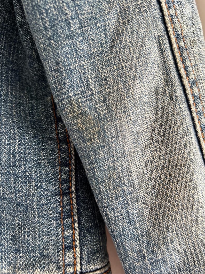 May include: Close-up of a denim jacket sleeve. The blue fabric shows a textured weave with visible stitching in a contrasting orange-brown thread. The jacket appears worn, with subtle fading.