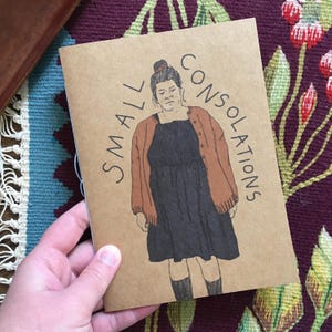 Può includere: Un libretto di carta marrone con il testo "SMALL CONSOLATIONS" stampato sulla copertina. La copertina presenta un'illustrazione disegnata a mano di una persona che indossa un cardigan marrone e un vestito nero.