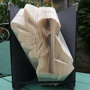 Peut inclure: Une sculpture d'art de livre représentant un oiseau, plié à partir des pages d'un livre, présentée dans un cadre noir avec des accents argentés.